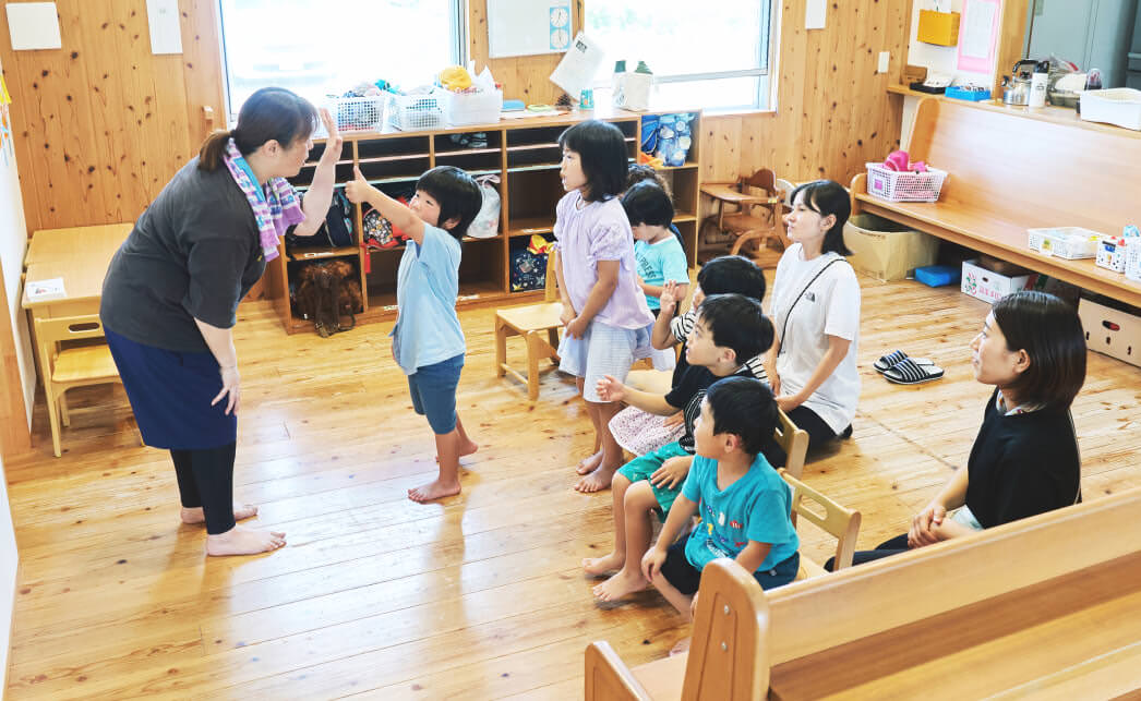 教室で、6人の子供が椅子に座っており、子供の前にいる保育士が遊びを通じた学習を行っている。補助で2人の保育士が子供の背後に座っている。